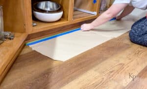 Putting masking paper on the floor around the perimeter of base cabinets in preparation to paint.